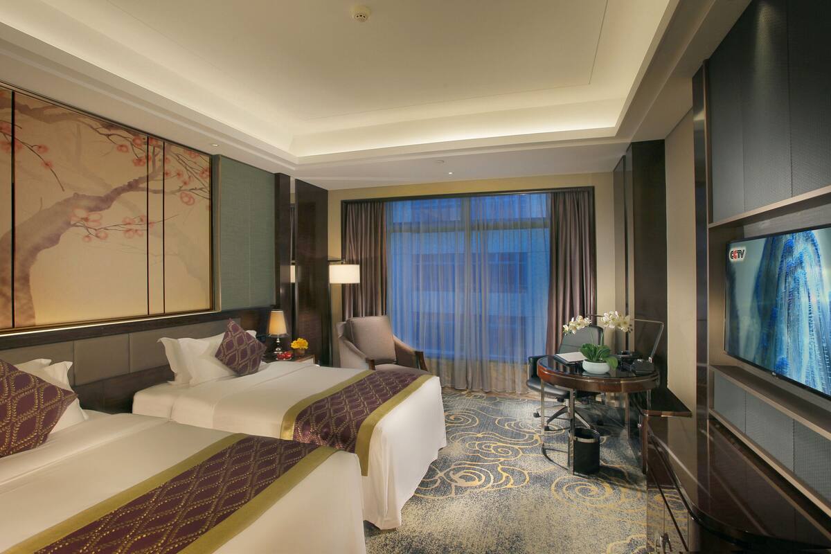 A hotel room with two beds, a desk with a laptop, a TV, and a large window with curtains.