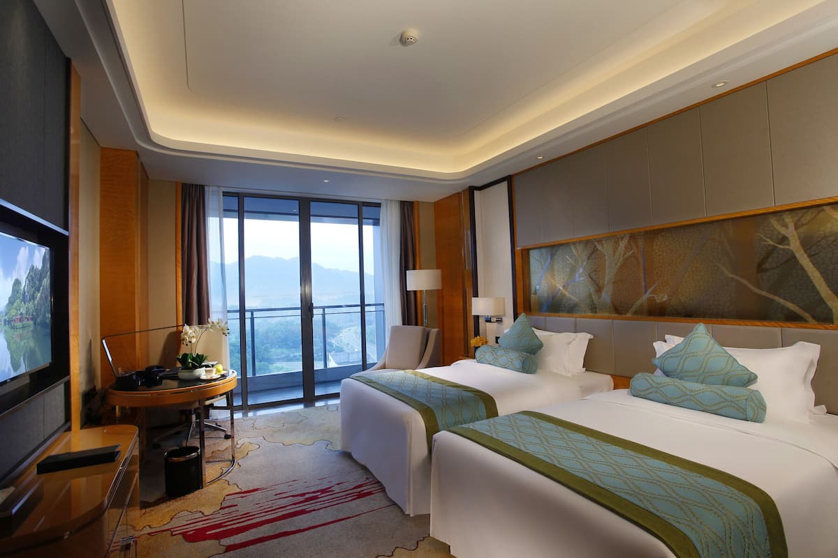 A hotel room with two beds, a TV, a desk, and a large window offering a mountain view.