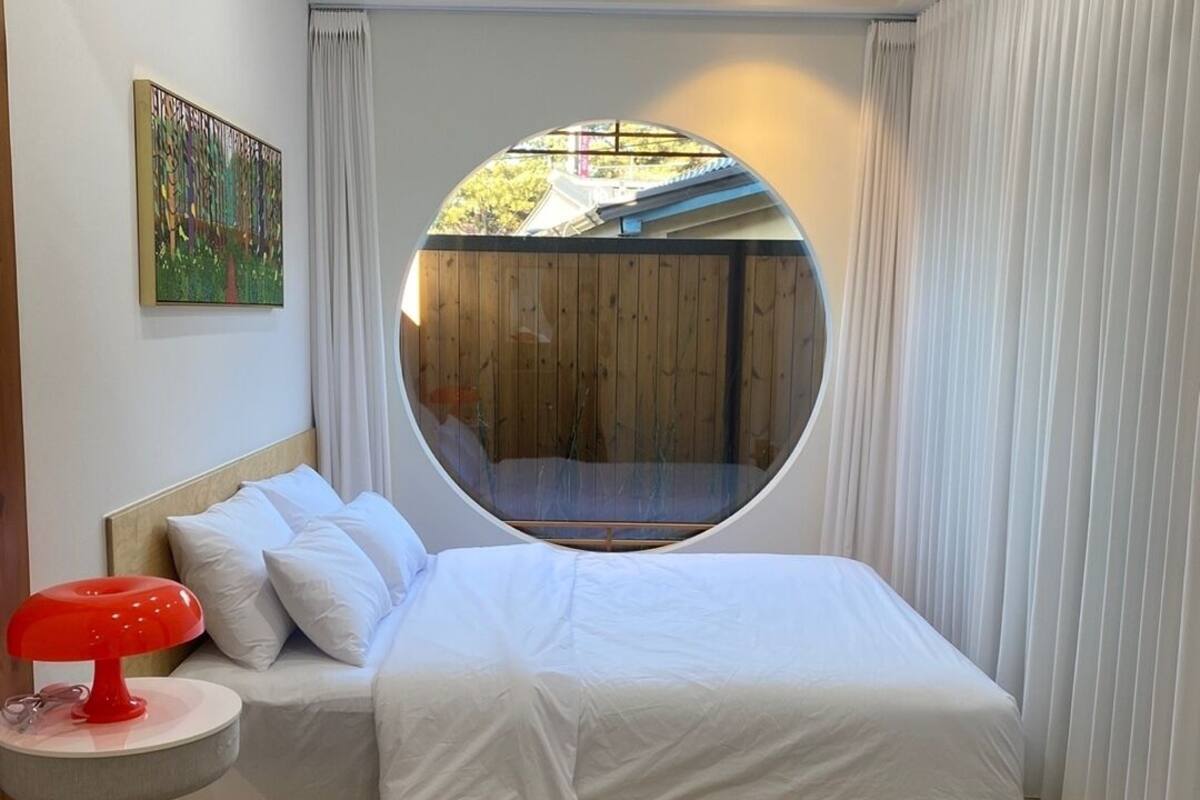 A bedroom with a circular window, a bed, a small table with a red lamp, and a painting on the wall.