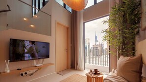 Un salon moderne doté d’un téléviseur à écran plat, d’une grande fenêtre offrant une vue sur la ville, d’un canapé confortable et d’une petite table d’appoint en bois.