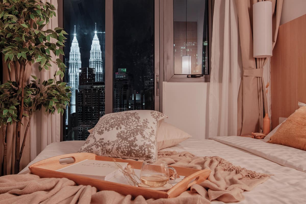 A bedroom with a large window, a bed with a wooden tray, a plant, and a view of the Petronas Towers at night.