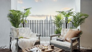 Un balcon avec deux fauteuils blancs dotés de coussins, une table en bois sur laquelle se trouve un plateau avec du thé et des en-cas, et une vue sur un paysage urbain dominé par une tour imposante.