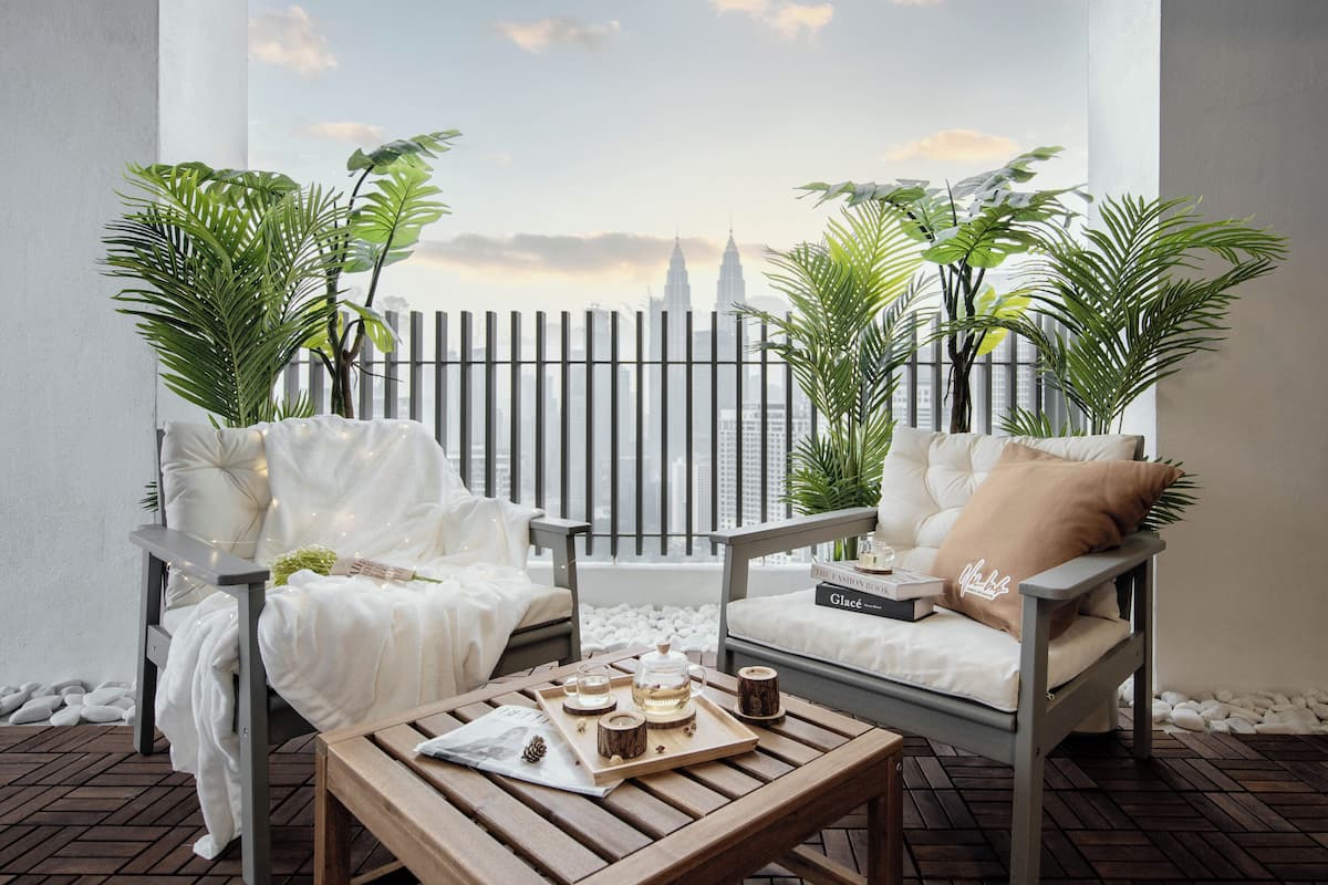A balcony with two white cushioned chairs, a wooden table with a tray of tea and snacks, and a view of a cityscape with a prominent tower.