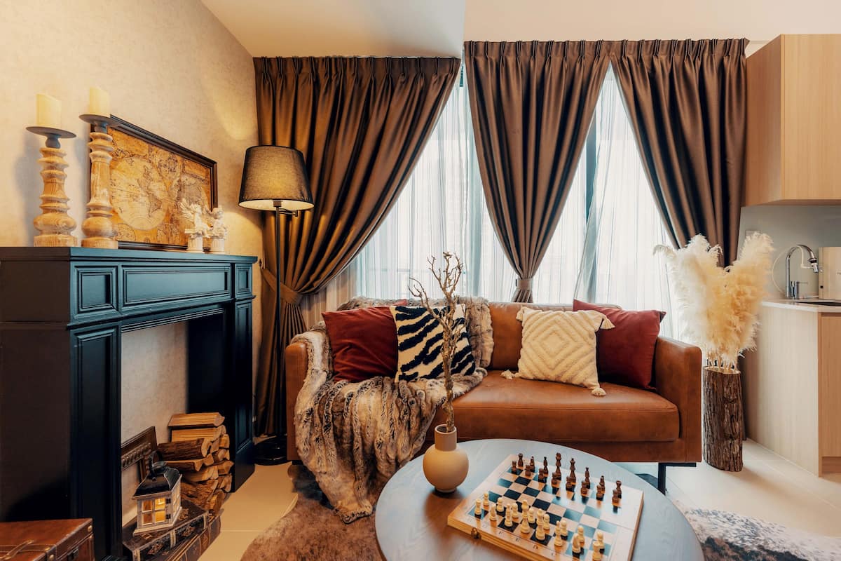 A cozy living room with a fireplace, a sofa, a coffee table with a chessboard, and a large window with curtains.