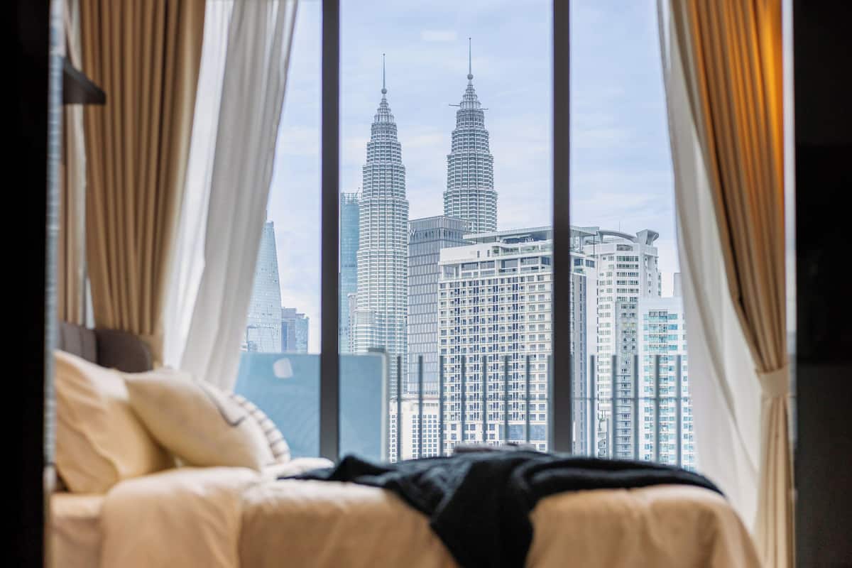 A hotel room with a bed, curtains, and a view of the Petronas Towers.