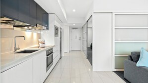 Une cuisine moderne avec des meubles blancs, des appareils en acier inoxydable et un canapé gris agrémenté d’un coussin bleu dans le salon attenant.
