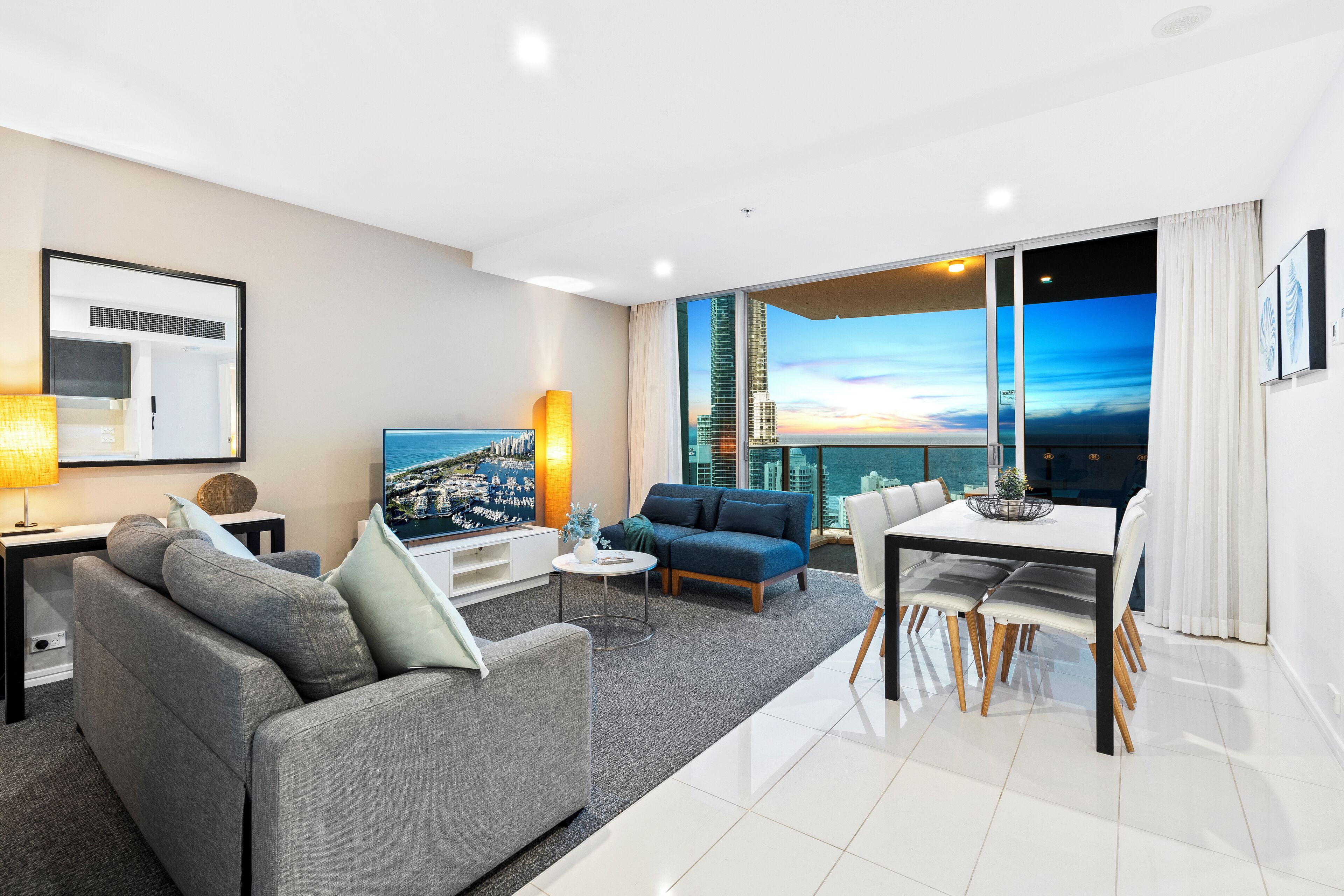 A modern living room with a sofa, coffee table, and dining area. A large window offers a city view.