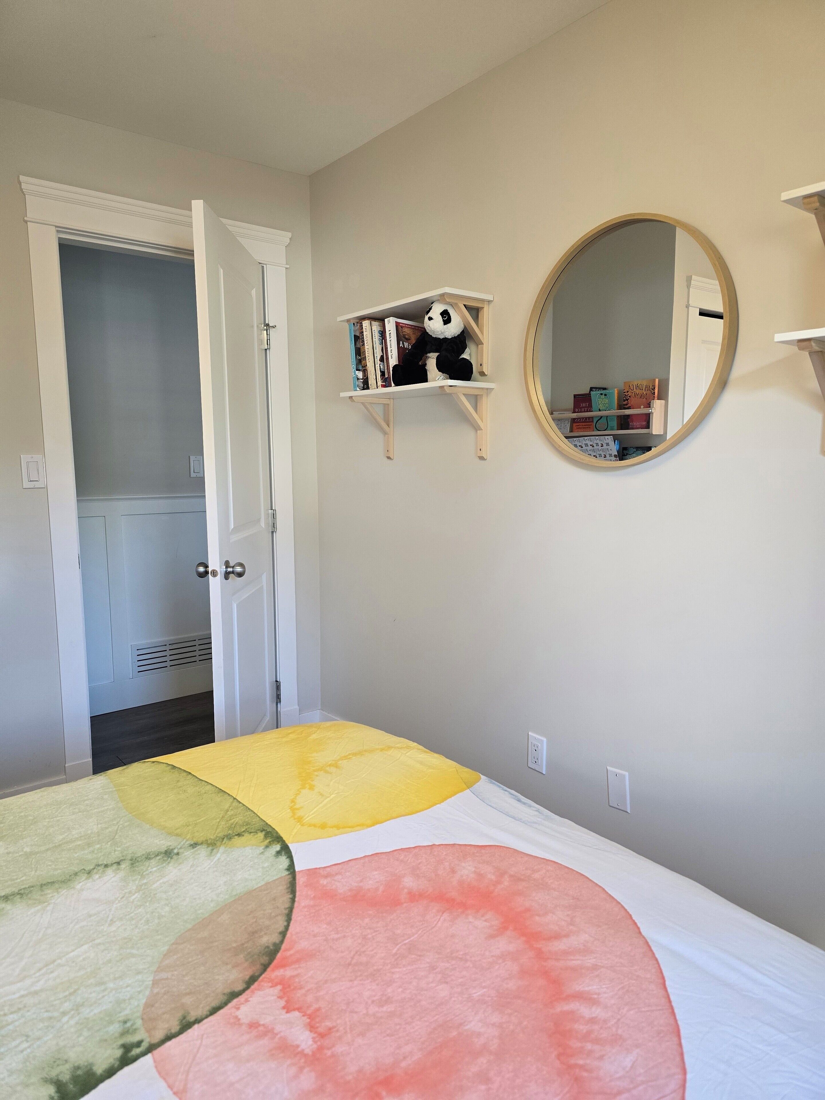 A bedroom with a bed, a shelf with books and a stuffed panda, a mirror, and a door leading to another room.