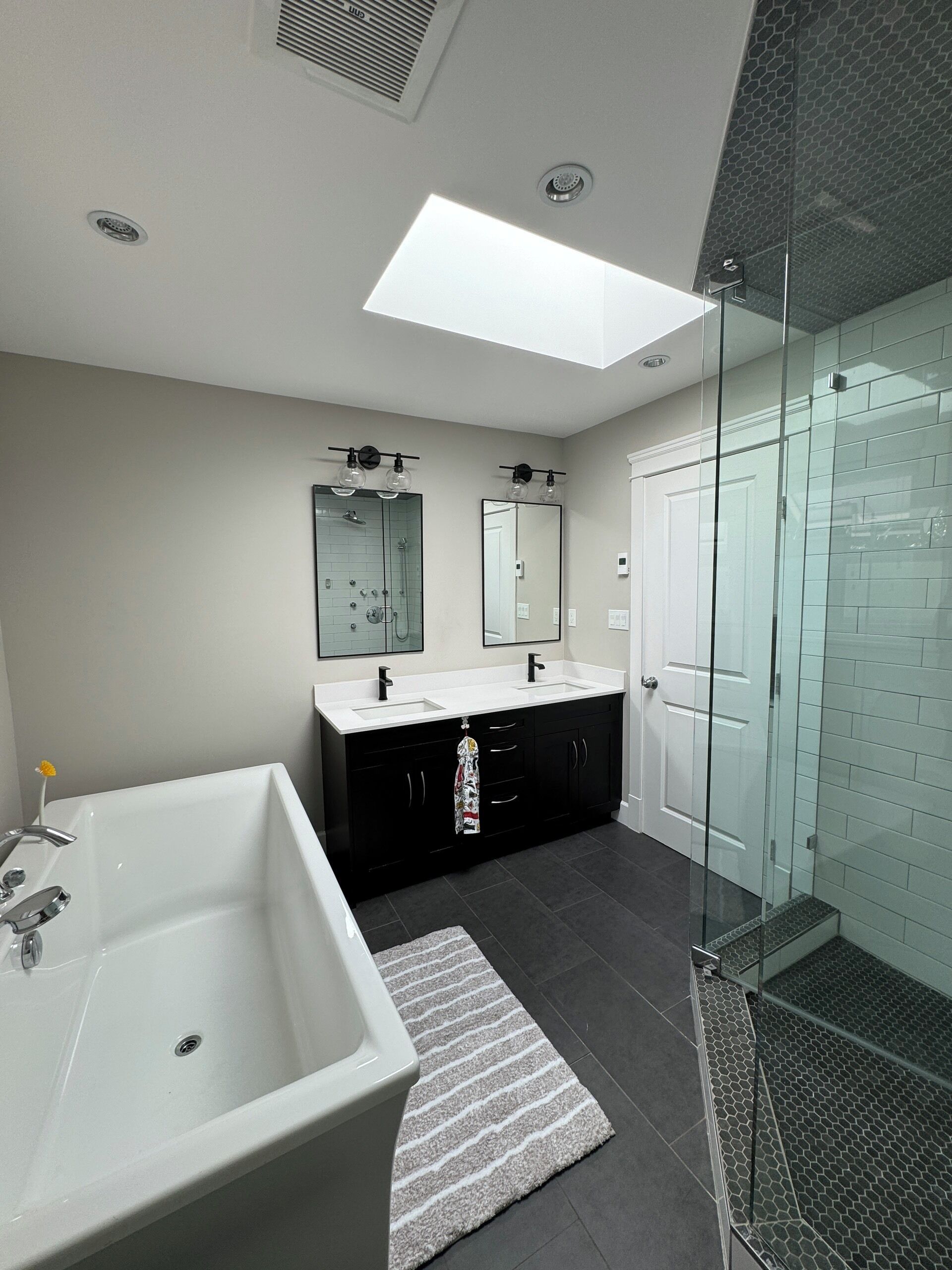 A modern bathroom with a large bathtub, two sinks, a glass shower enclosure, and a skylight.