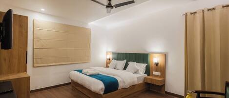 A hotel room with a wooden bed, a ceiling fan, a bedside table, a chair, and a glass table with a bottle.