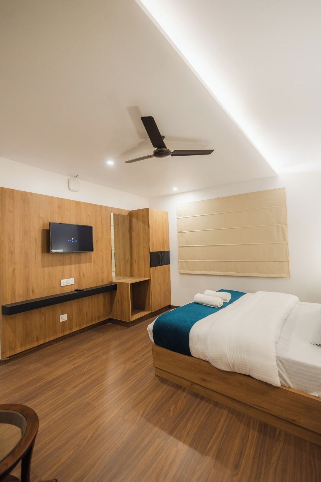 A modern hotel room with a bed, wooden wardrobe, ceiling fan, and a mounted TV.