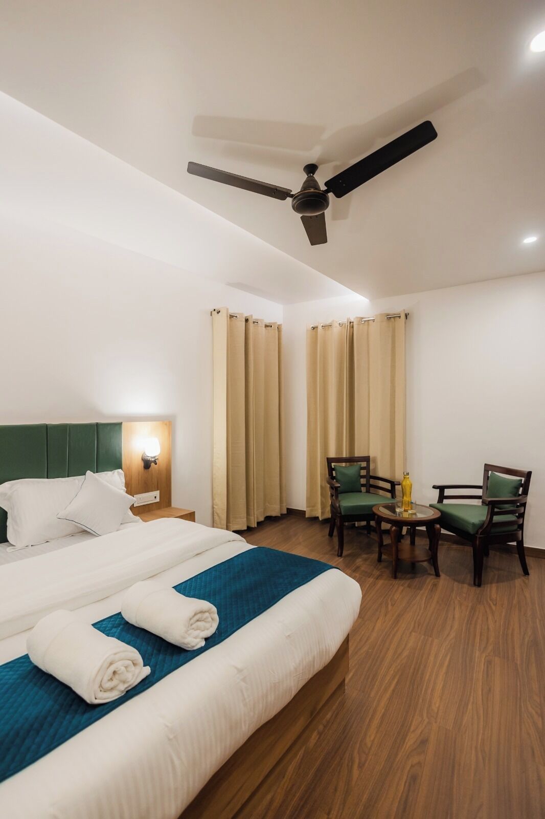 Una habitación de hotel con cama, dos sillones, una mesa, ventilador de techo y cortinas.