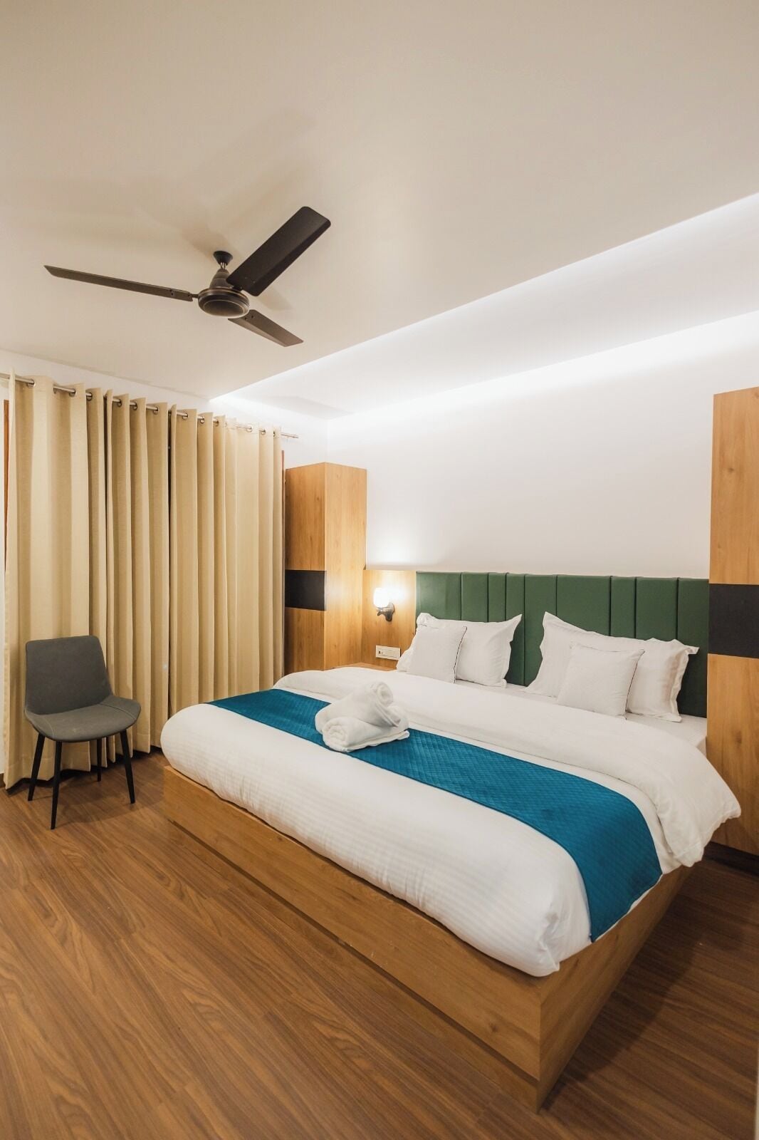 A hotel room with a wooden bed, a green headboard, a ceiling fan, a chair, and a window with curtains.