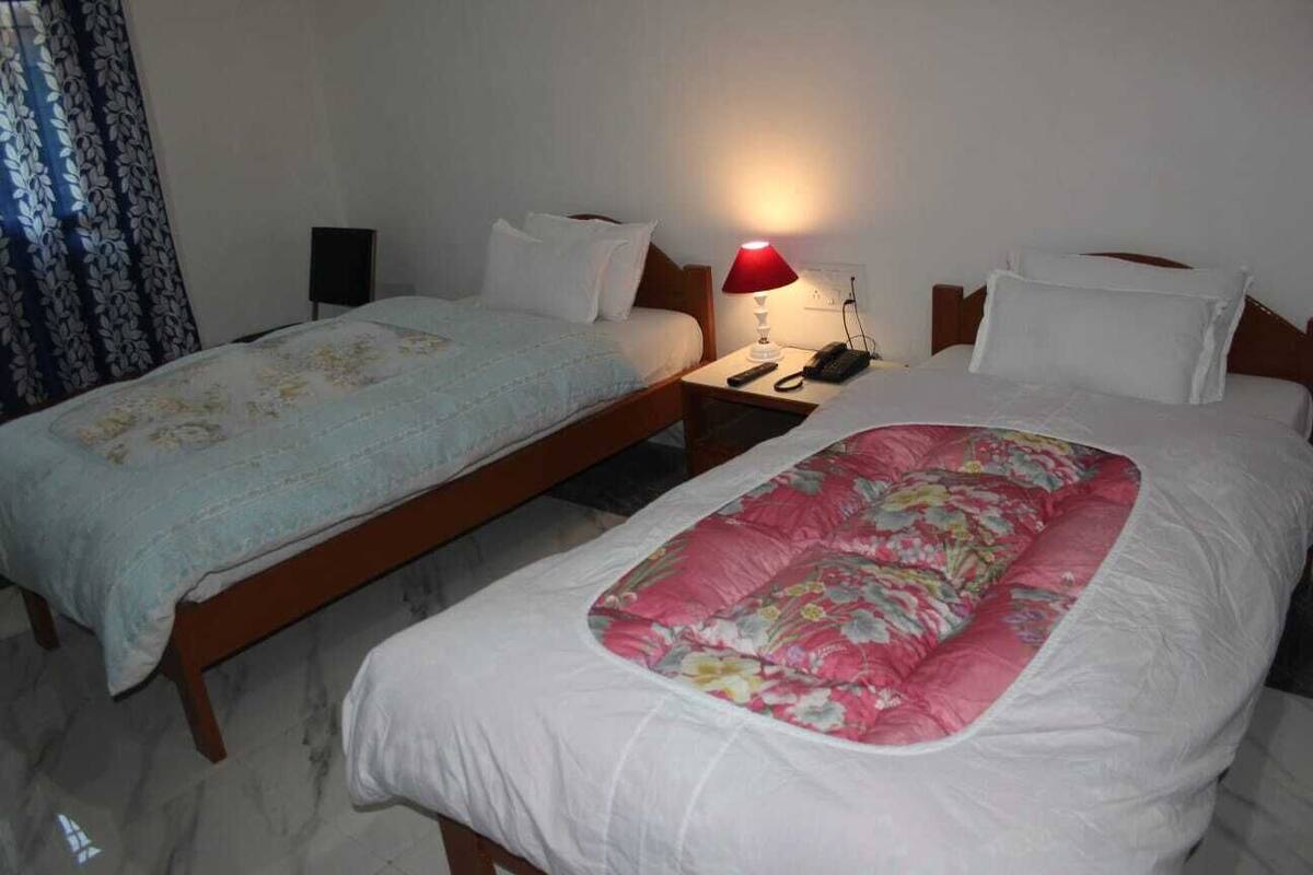 A hotel room with two single beds, a nightstand with a lamp, a phone, and a wall socket.