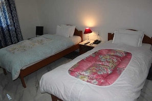 A hotel room with two single beds, a nightstand with a lamp, a phone, and a wall socket.