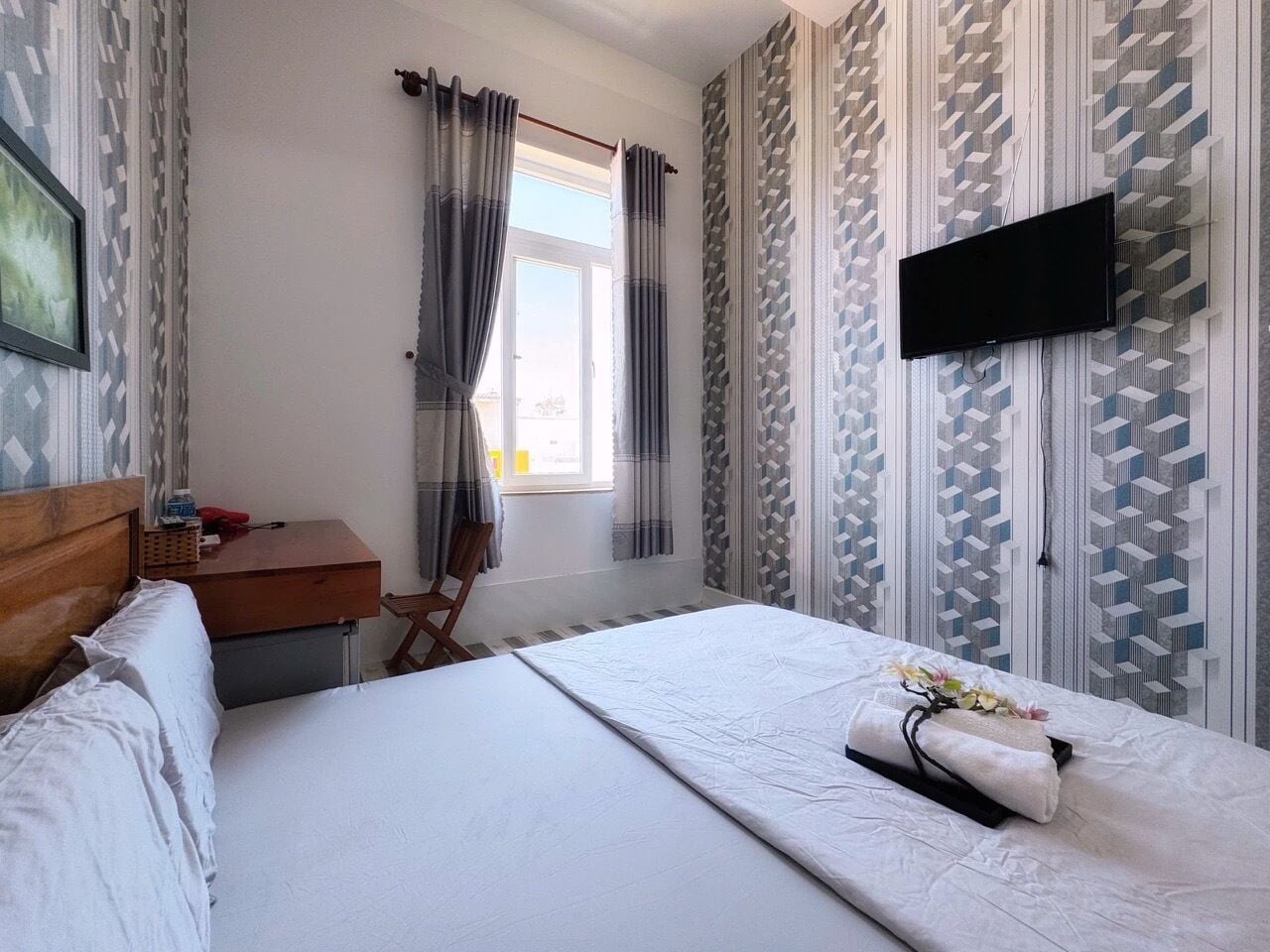 A hotel room with a bed, a wooden nightstand, a chair, a TV mounted on the wall, and patterned curtains.