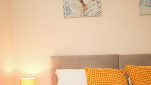 A neatly made bed with white and orange pillows, a white throw blanket, and a bedside lamp on a wooden nightstand.