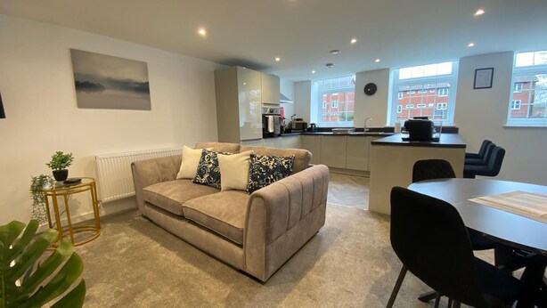 Een moderne woonkamer met een beige bank, een klein bijzettafeltje met een plant, en een eethoek met een tafel en stoelen.