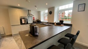 Een moderne keuken met een ingebouwd eiland, barstoelen, een koffieapparaat en uitzicht op buiten door grote ramen.