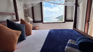 A bed with a white and blue blanket, multiple pillows in various colors, a nightstand with a lamp, and a window with a view of the ocean.