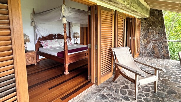 A wooden four-poster bed with a canopy, a stone wall, and a lounge chair outside a room with wooden shutters.