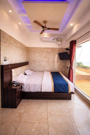 A bedroom with a bed, a ceiling fan, an air conditioning unit, a TV, and a window with a view. - Blue Pearl Beach Guest House (Bhatkal)