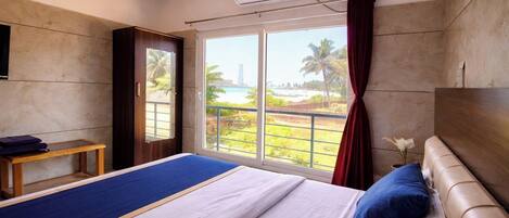 Quarto de hotel com cama, uma pequena mesa, televisão e janela com vista para a praia e palmeiras.