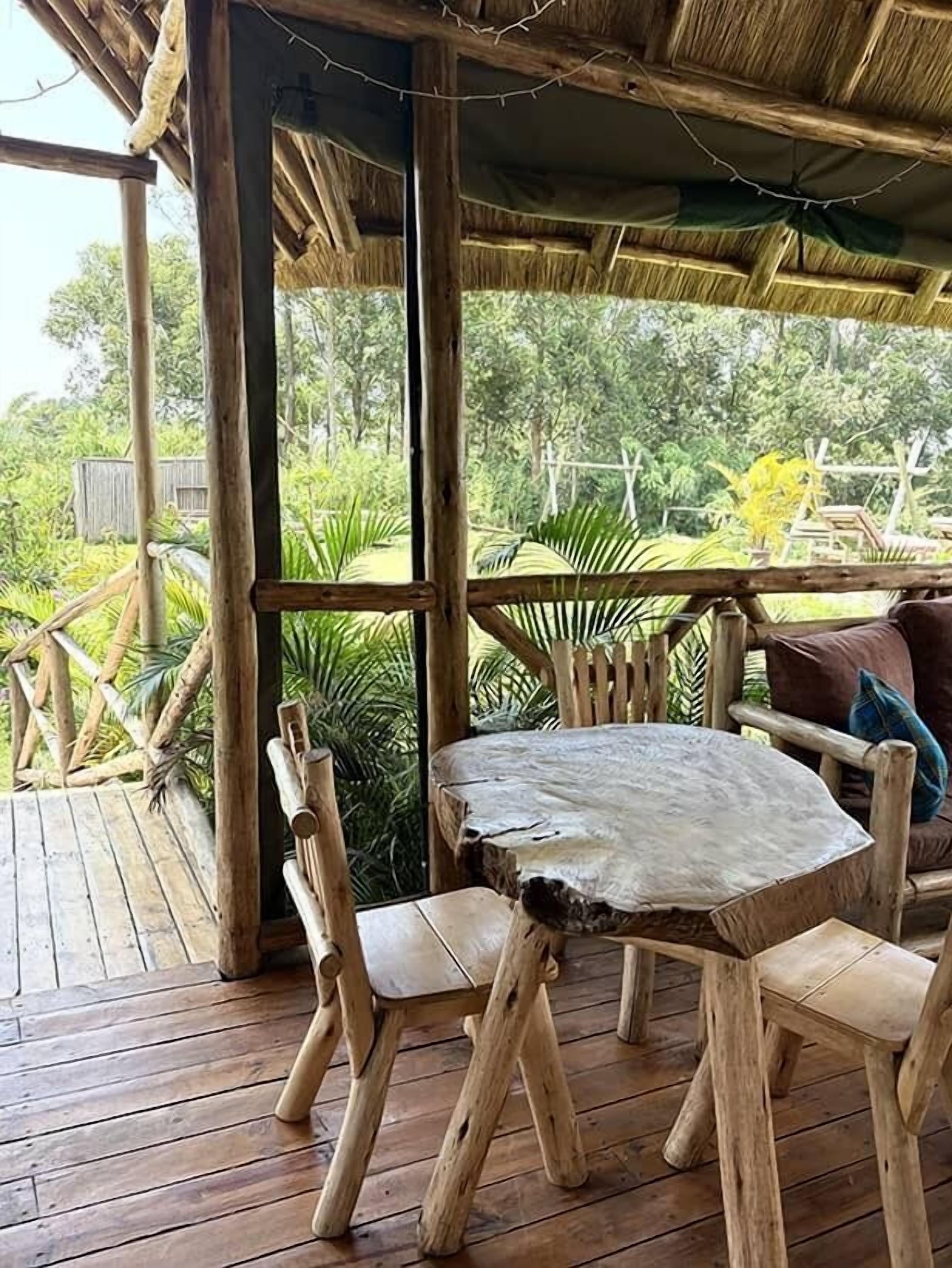 Una terraza de madera al aire libre con una mesa redonda y sillas, rodeada de exuberante vegetación y una estructura de techo de paja.