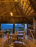 Ein rustikaler Essbereich aus Holz mit Tischen und Stühlen, einem Reetdach und Blick auf die Landschaft.