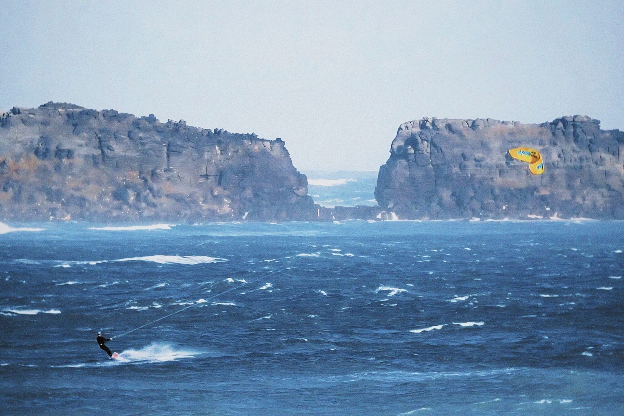 A person kiteboarding in a body of water with rocky cliffs in the background.