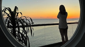 Una persona de pie junto a una ventana con vista a una puesta de sol sobre el océano.