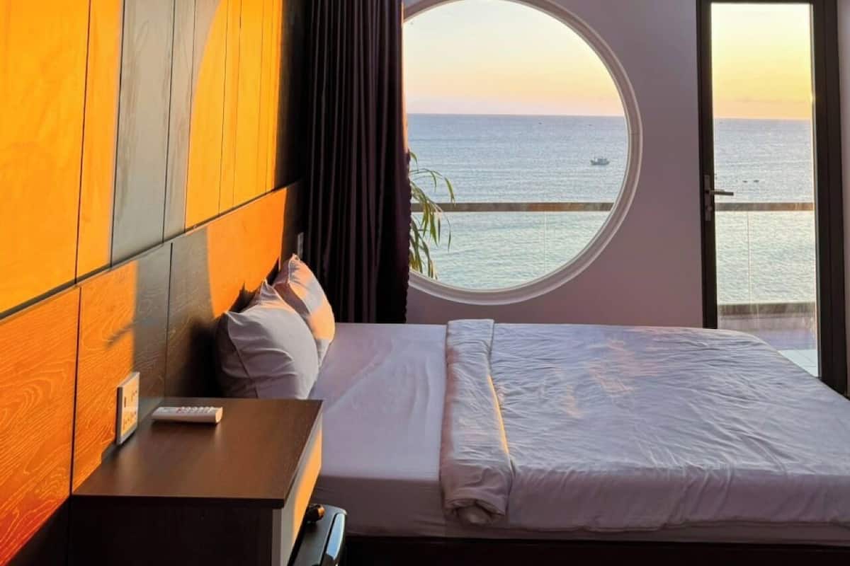 Un dormitorio con una cama grande, una mesita de noche, un ventanal redondo con vistas al mar y una puerta al exterior.