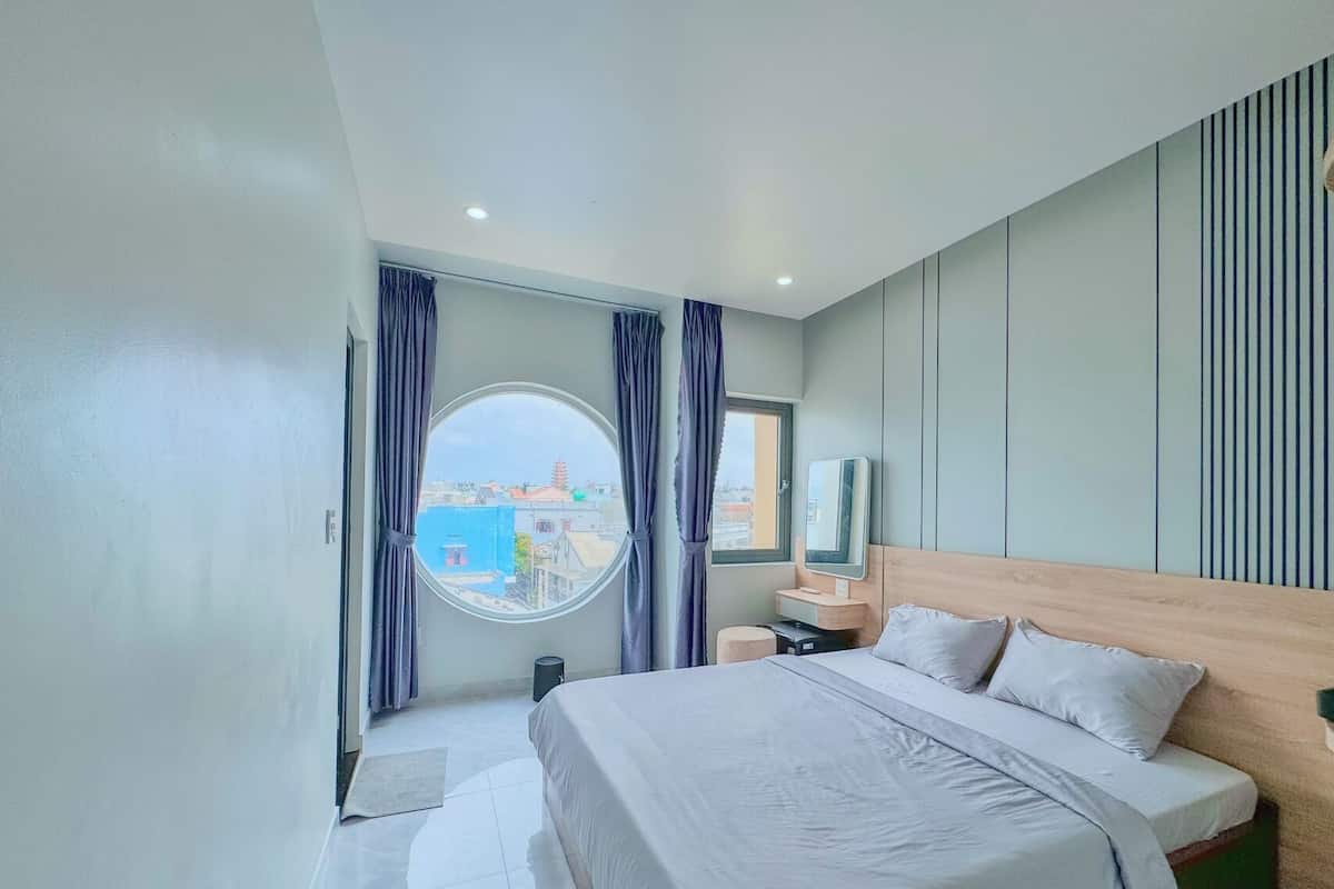 Habitación de hotel moderna con una cama grande, un ventanal redondo y vistas a edificios.
