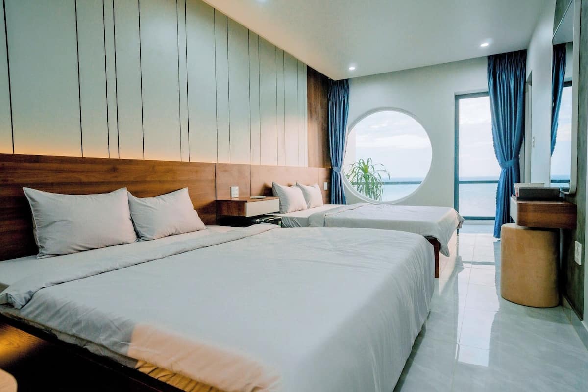 Una habitación de hotel moderna con dos camas, un cabecero de madera, un ventanal redondo y vista al mar.