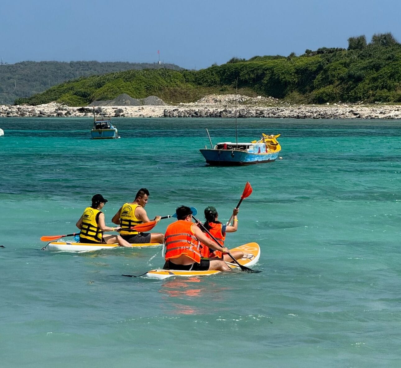 Een groep mensen kajakt op een heldere turquoise zee, met rotsachtige oevers en begroeiing op de achtergrond.