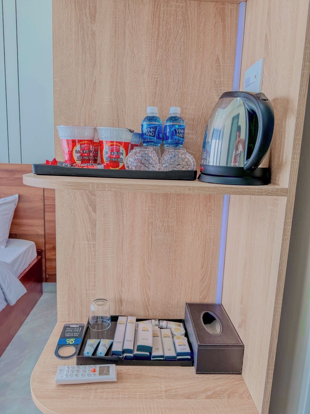 A hotel room with a wooden shelf holding toiletries, a kettle, and a mirror. Below, there's a tray with a remote control, a glass, and a box of tissues.
