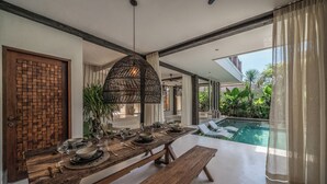 A dining area with a long wooden table set for four, a hanging light fixture, and a view of a swimming pool. - Casa Galicia (Canggu)