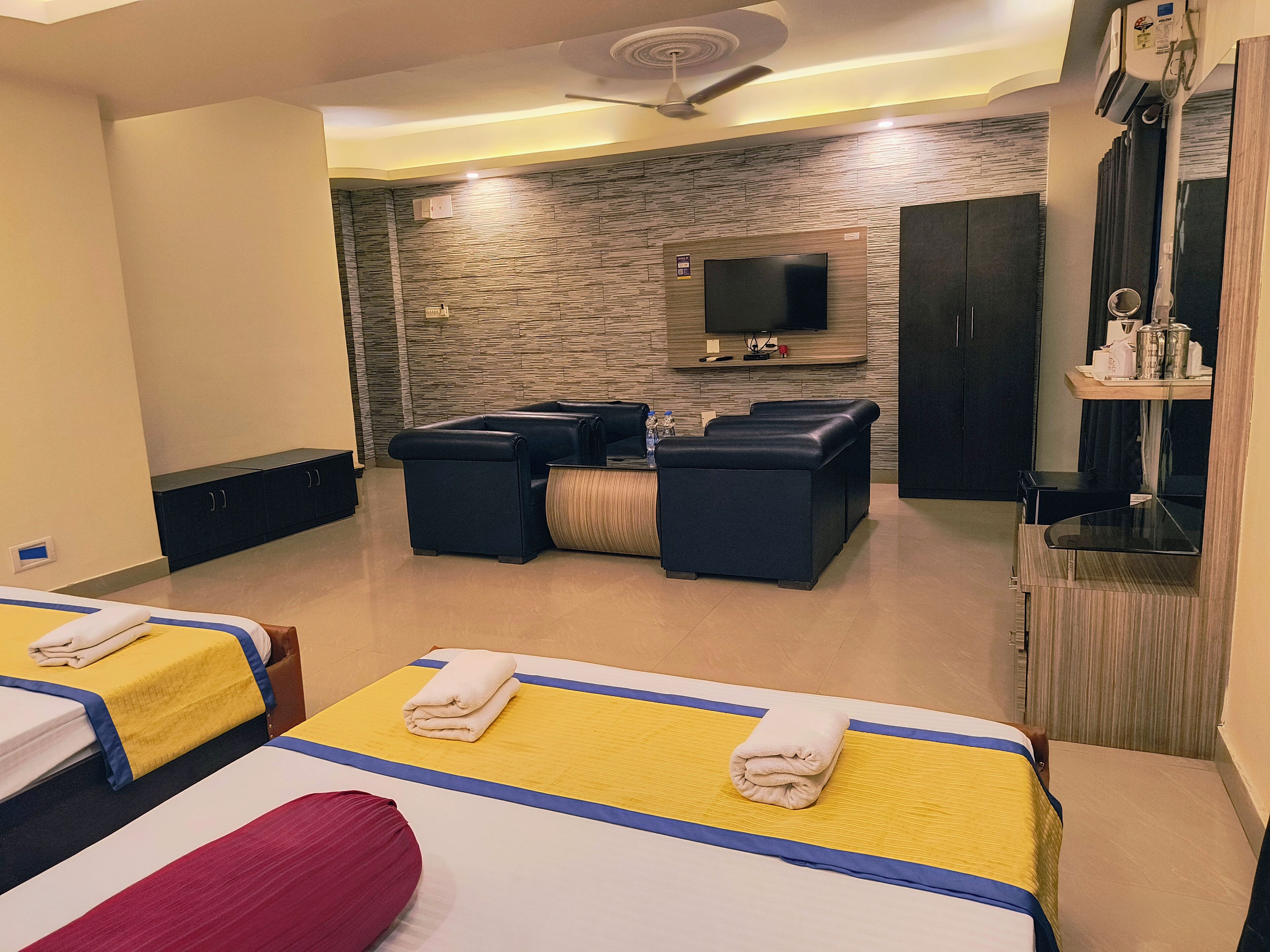 A hotel room with two beds, a seating area with sofas, a flat-screen TV mounted on the wall, and a ceiling fan.