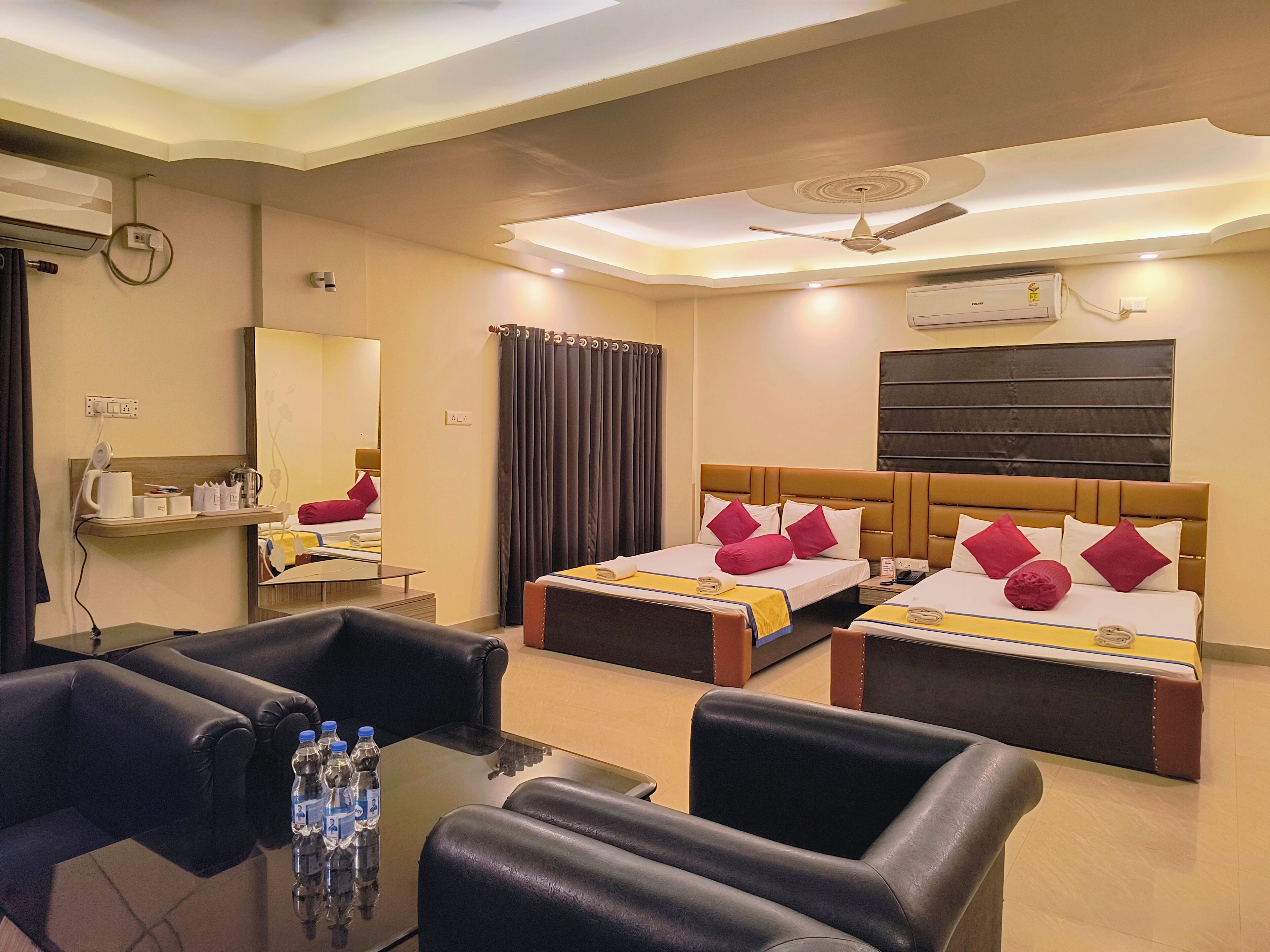 Habitación de hotel con dos camas, una zona de estar con sillas, una mesa con botellas de agua, un espejo y un equipo de aire acondicionado.
