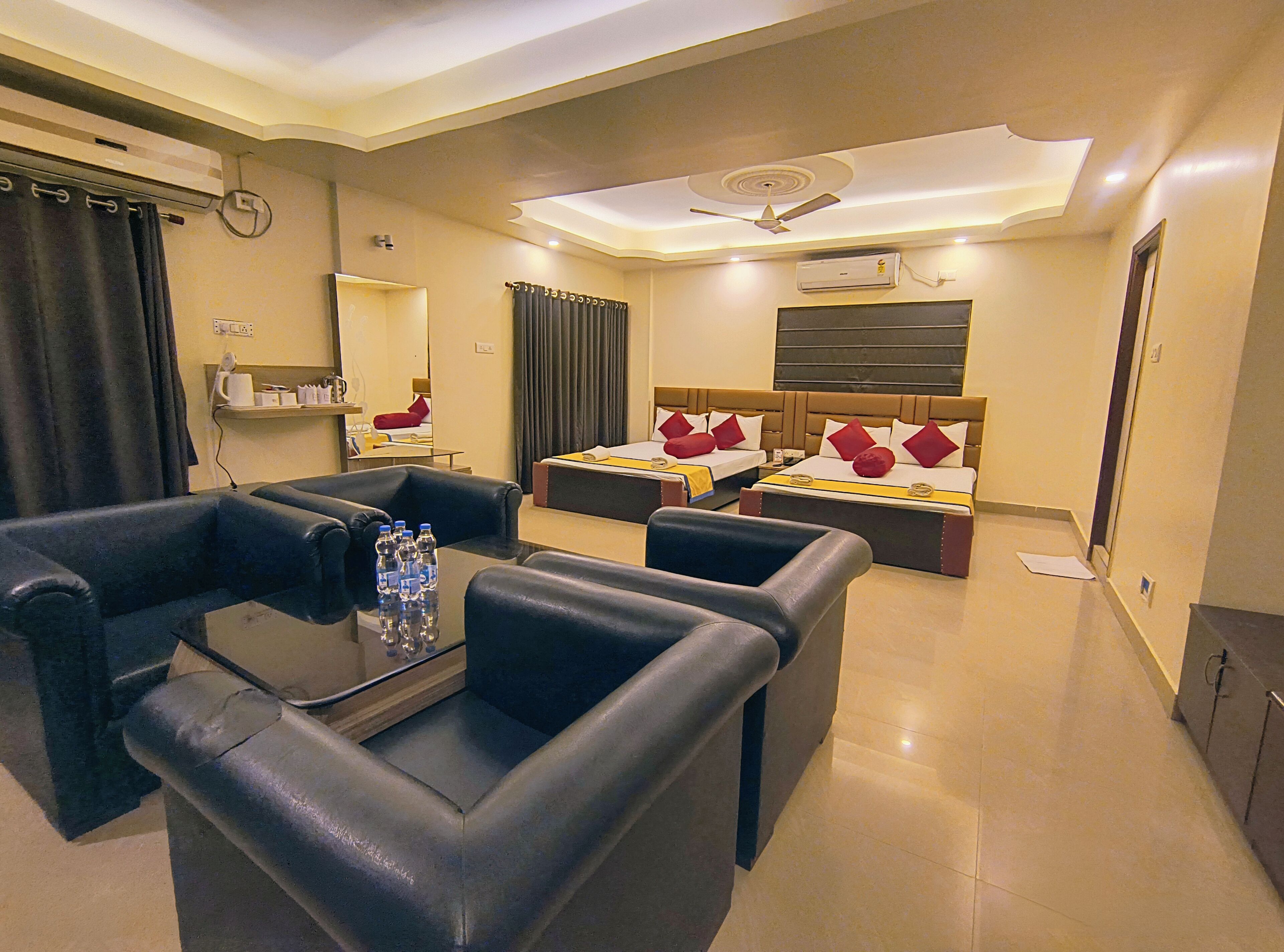 A hotel room with a sofa set, a small table with water bottles, a bed with red pillows, and a ceiling fan.