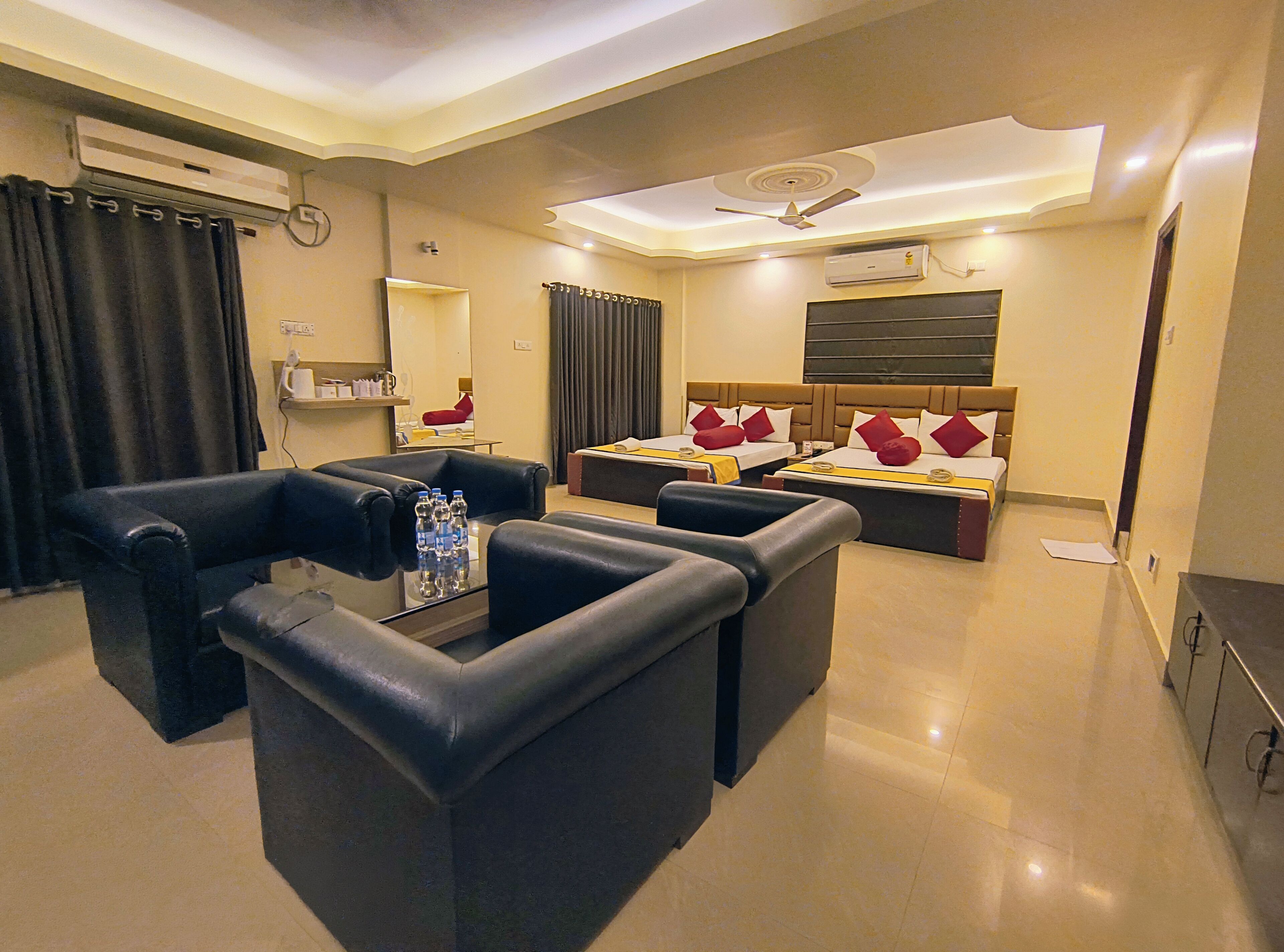 A hotel room with a sofa set, a bed with red pillows, a ceiling fan, and an air conditioning unit.