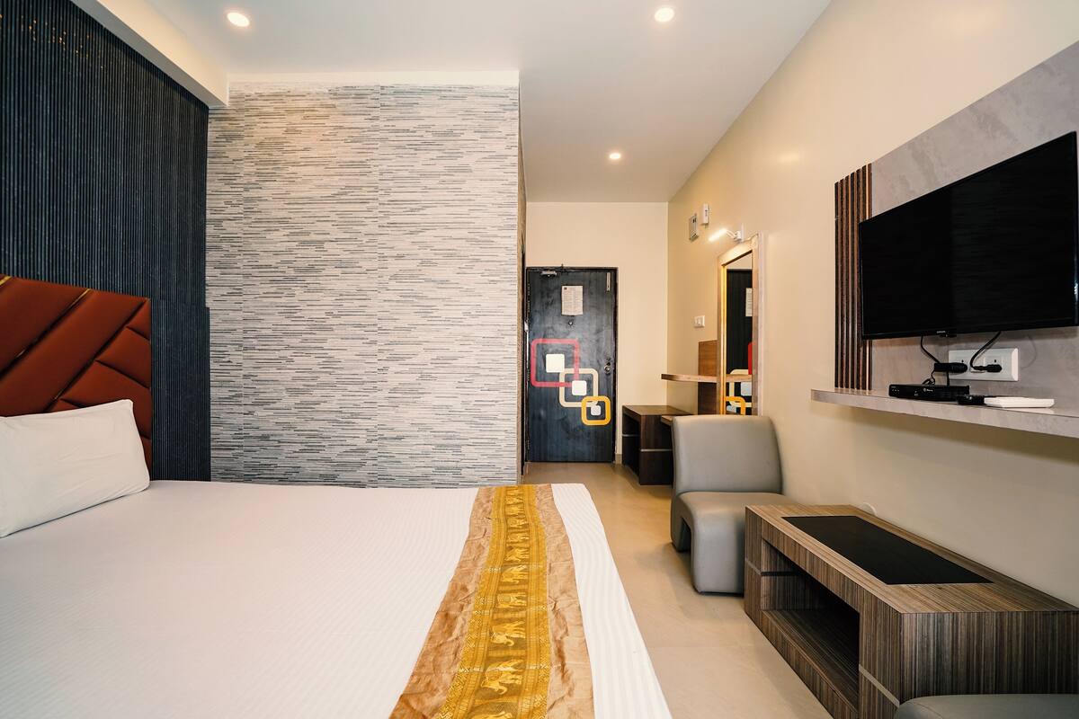 Una habitación de hotel moderna con una cama grande, un televisor de pantalla plana en la pared, un espacio de descanso con un banco y una puerta con un símbolo cuadrado rojo.