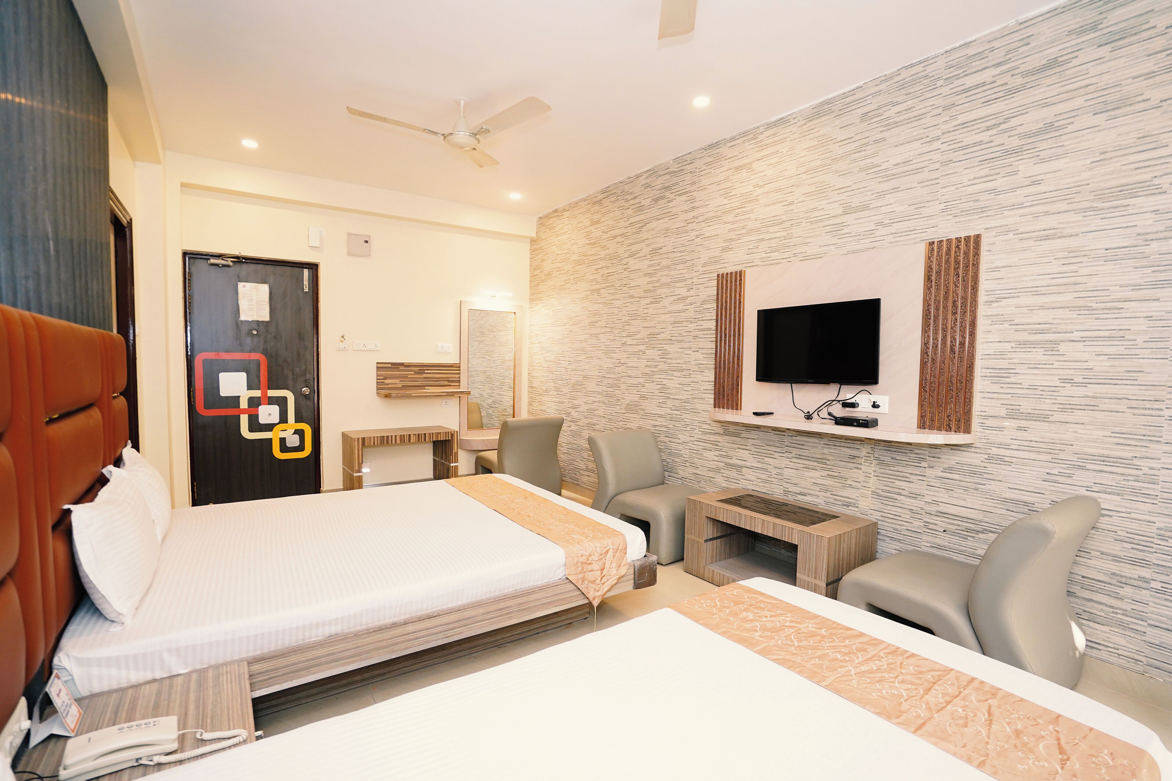 Ein Hotelzimmer mit Bett, Schreibtisch, Stuhl, Wandfernseher und Deckenventilator.