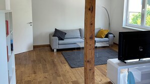 Una sala moderna con una viga de madera, un sofá gris, un mueble de TV blanco y una ventana con vistas a la vegetación.