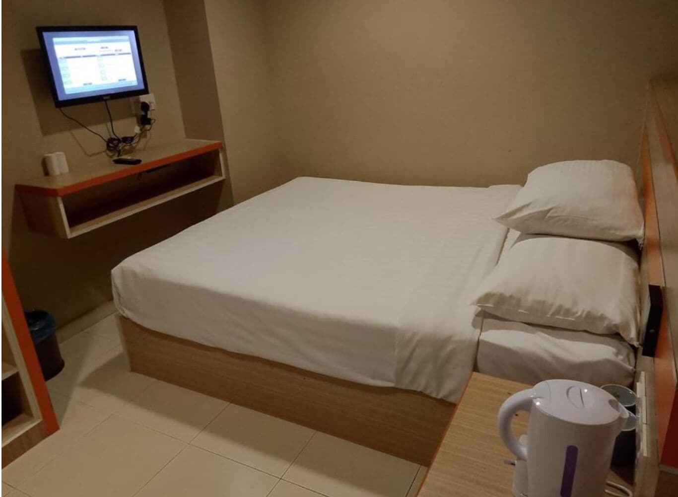 Ein kleines, ordentliches Schlafzimmer mit einem Bett, einem Regal mit einem Computermonitor und einem Wasserkocher auf dem Nachttisch.