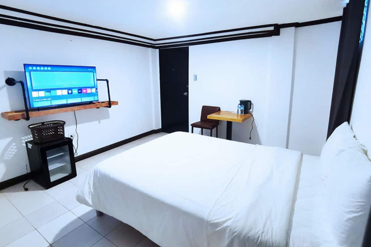 Quarto de hotel com cama, escrivaninha, cadeira, televisão fixada na parede e um pequeno armário.