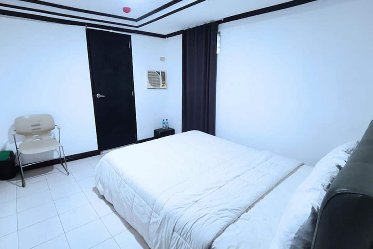 Quarto de hotel com uma cama, uma cadeira, uma mesinha e uma porta.