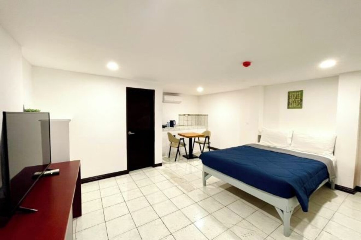 A bedroom with a bed, a desk, a chair, a small kitchenette, and a door.