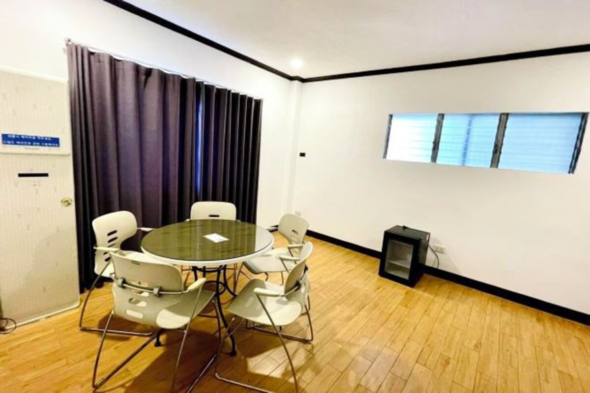 A conference room with a round glass table and white chairs, a black-framed window, and a wooden floor.