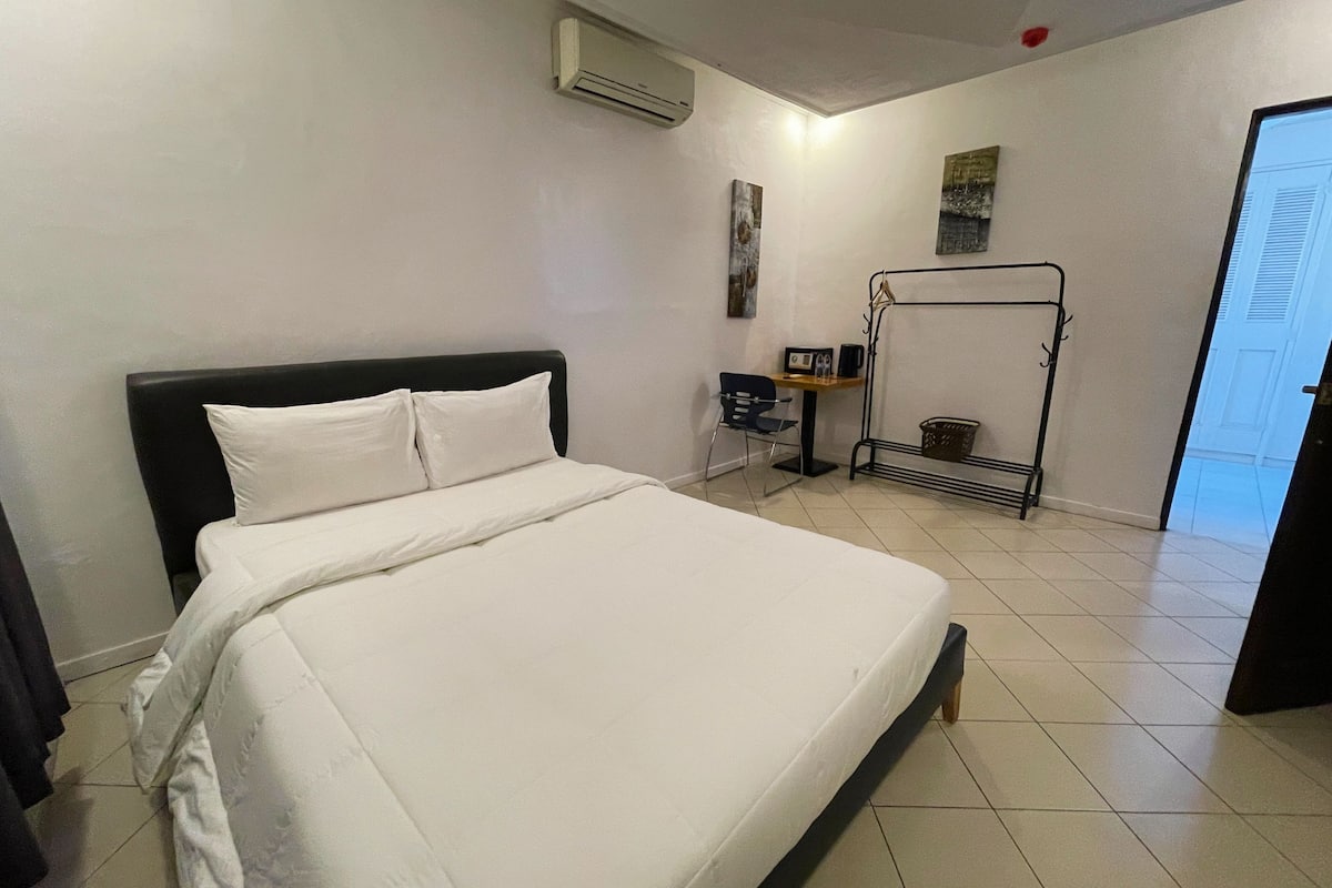 Quarto de hotel com uma cama grande, uma escrivaninha com cadeira, um cabideiro e um aparelho de ar condicionado.